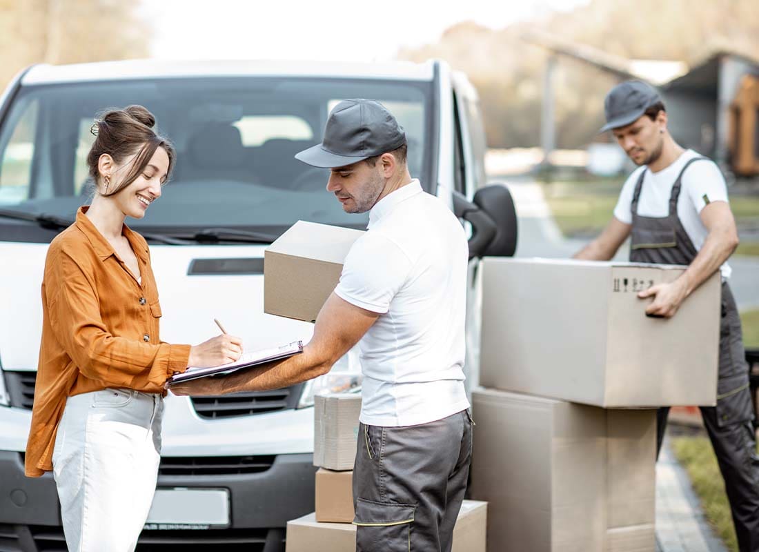 Moving Company Insurance - Moving Company Delivering Goods to a Young Woman by Cargo Van While She Is Signing Documents and Mover Unloads Cardboard Parcels in the Background